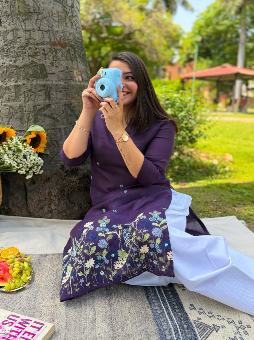 Woman in a purple dress with floral patterns sitting outdoors, taking a selfie.