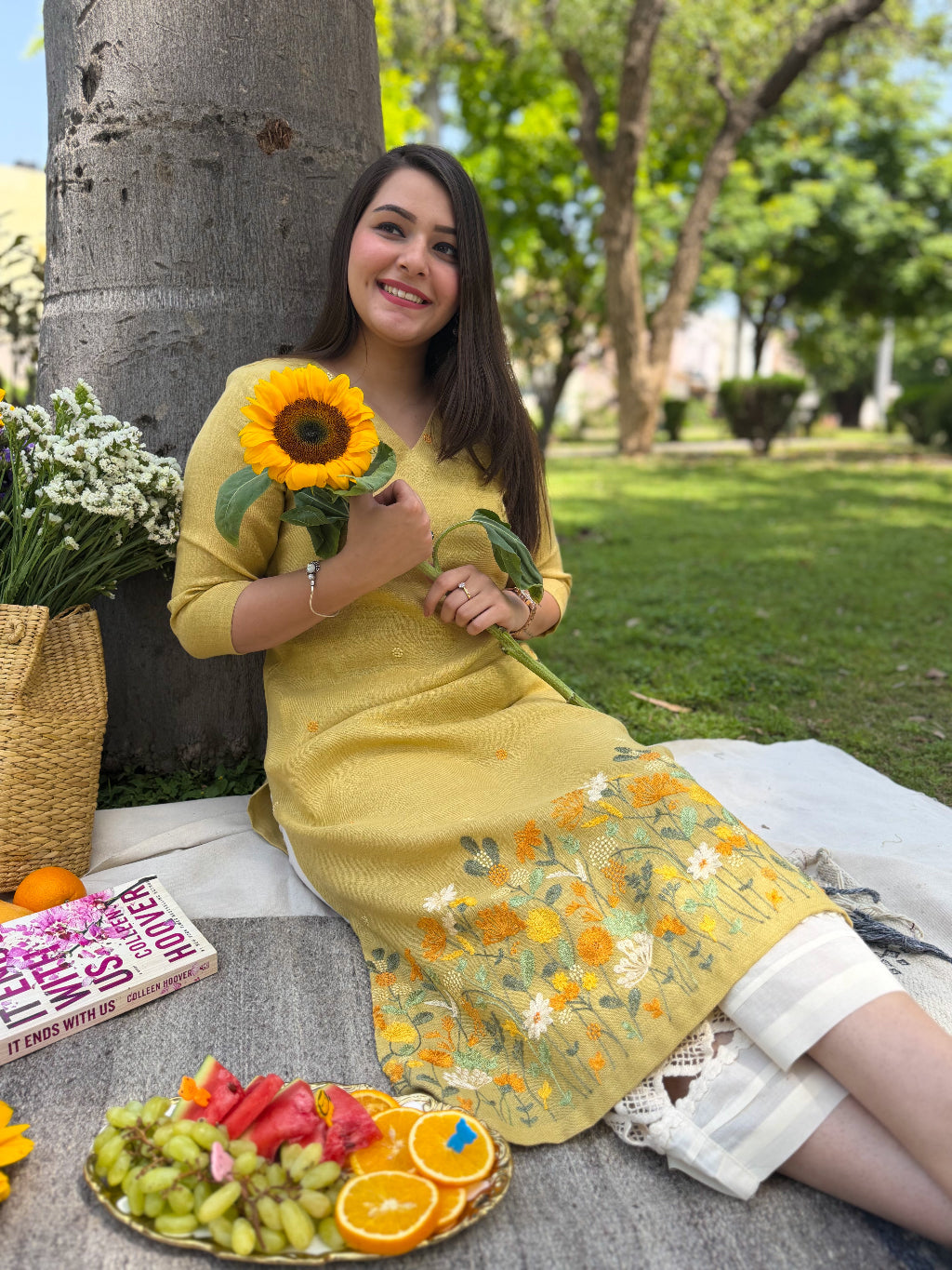 Woman in a yellow dress holding a sunflower in a park setting with a book and fruits on a table.