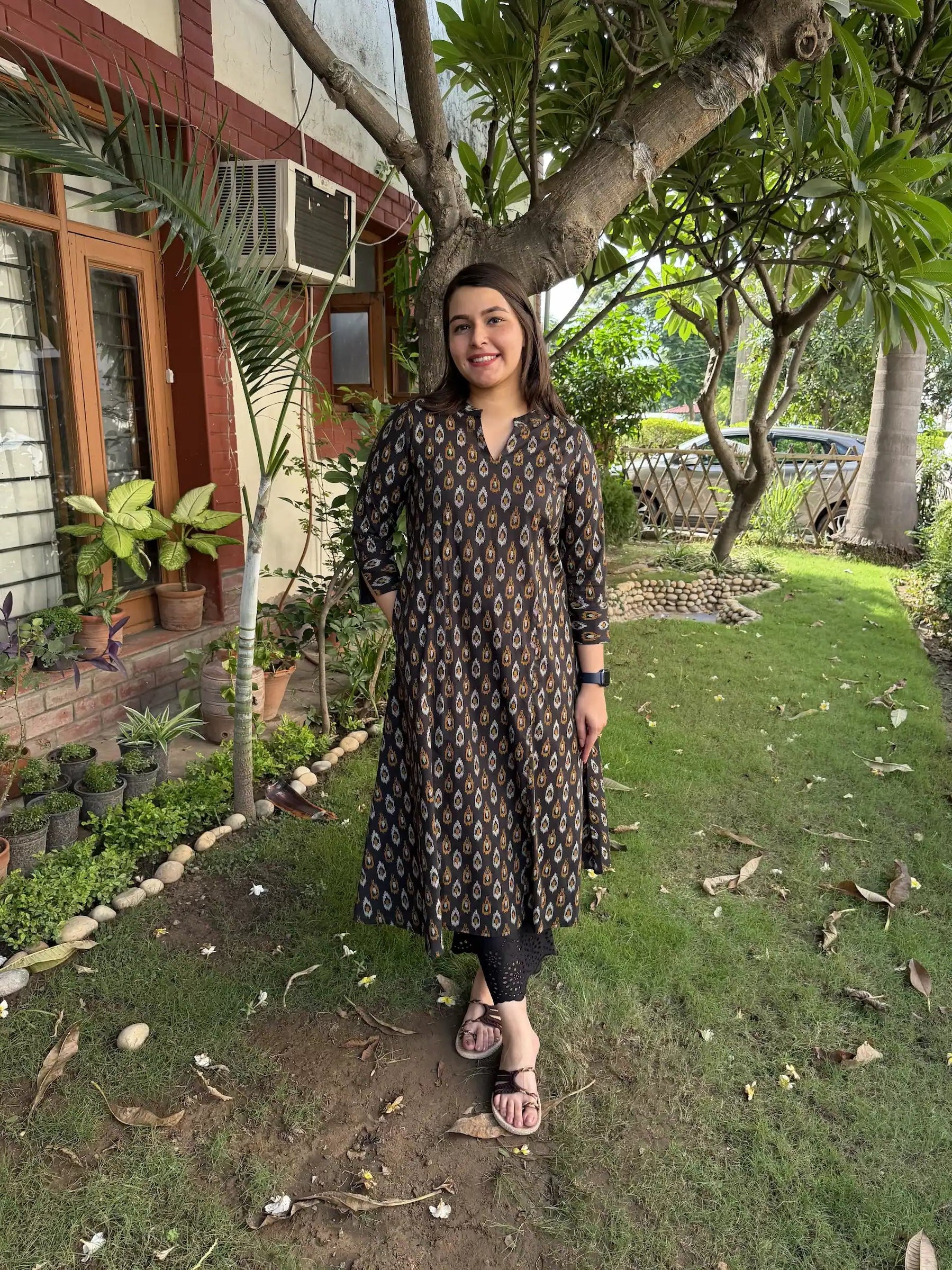 A woman posing outdoors in a dark brown patterned kurta with gold detailing, paired with black leggings and sandals, set against a background of green grass and trees.
