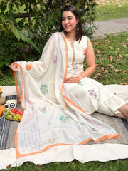 Woman in a white embroidered outfit sitting outdoors on a blanket with greenery around.