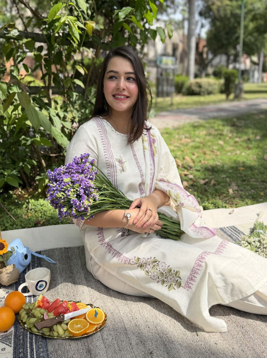 Woman in a white embroidered dress holding flowers, sitting outdoors with a plate of fruit and a cup.
