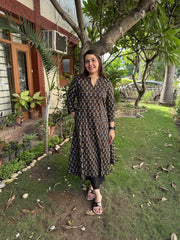 A woman posing outdoors in a dark brown patterned kurta with gold detailing, paired with black leggings and sandals, set against a background of green grass and trees.